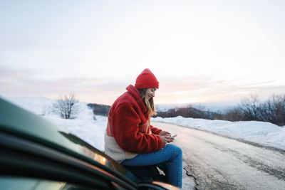 Frau sitzt auf Autodach und schaut auf Handy im Winter.jpg KI-generiertes Bild